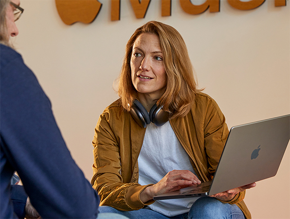 Dwie osoby z?zespo?u Sales and Business Development firmy Apple podczas rozmowy. Jedna osoba trzyma MacBooka i?siedzi naprzeciwko ?ciany z?logo Apple?Music.