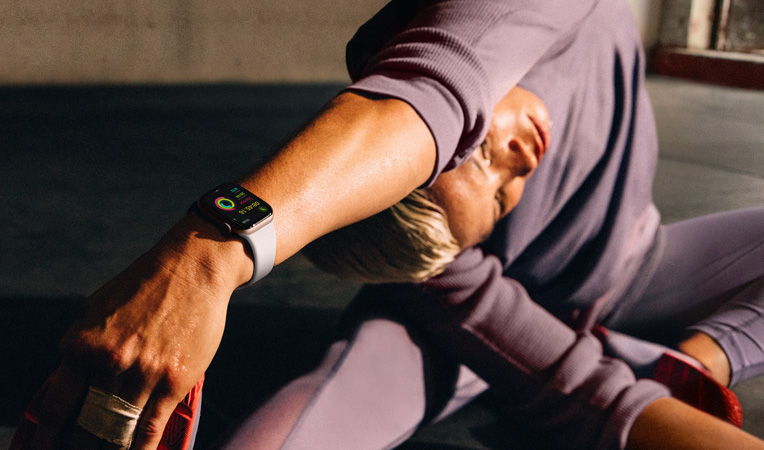 Apple Watch Series 10を裝著した女性が床に座り、手を足先に向けて伸ばしている。Apple Watchの畫(huà)面には、「ワークアウト」「クールダウン」「アクティビティ」とアクティビティリングが表示されている