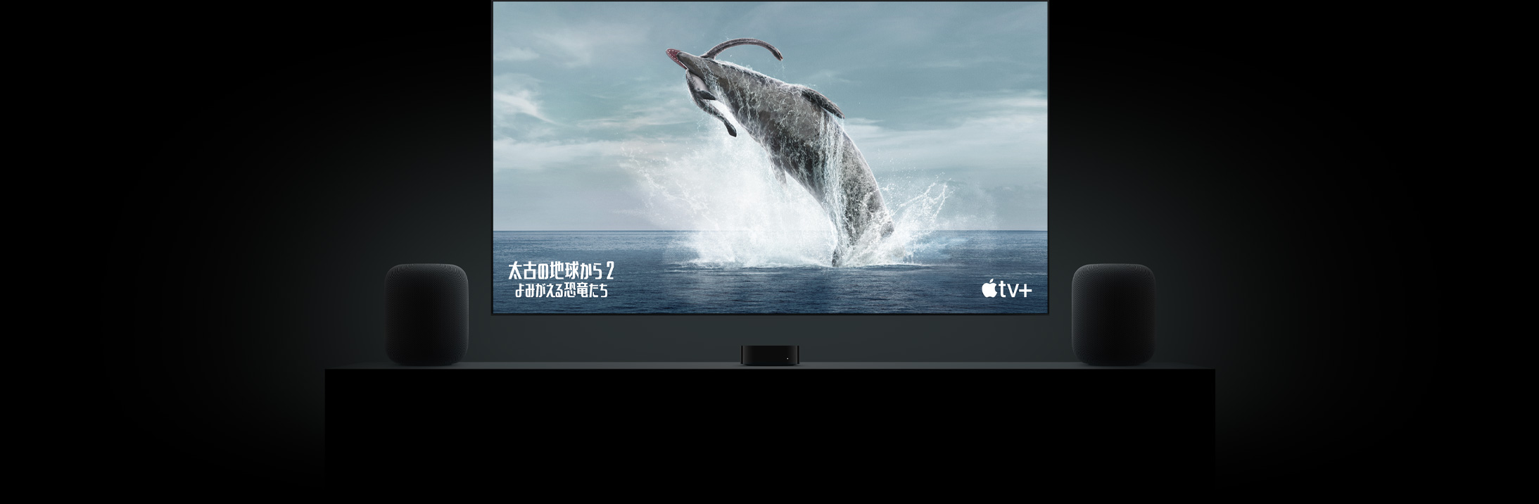 フラットテレビの大きなスクリーン上に「太古の地球から?よみがえる恐竜たち?」の恐竜の鮮明な映像が映し出されている。リビングルームのキャビネットの上に置かれた2臺(tái)のHomePodスピーカー。その間にApple TVが配置され、その上にテレビが掛けられている