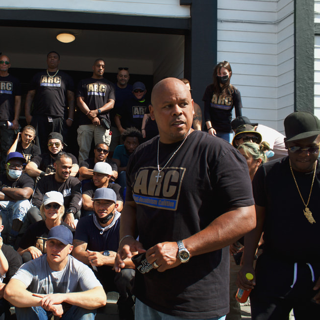 Orang-orang berkumpul di sekeliling Sam Lewis dalam Community Garden Day Anti-Recidivism Coalition di Magnolia Transitional Housing. 