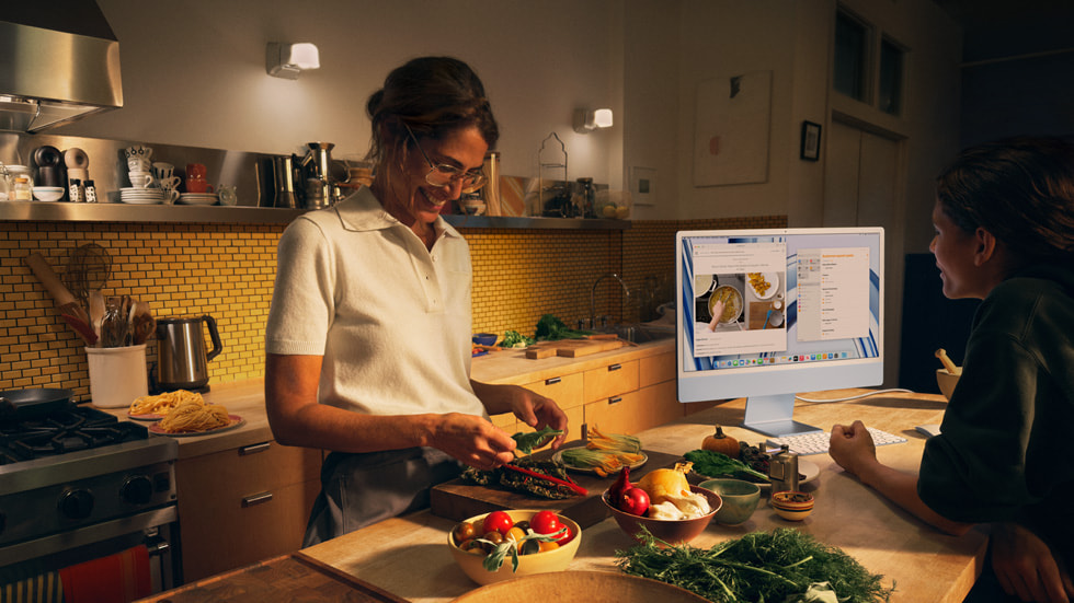 Orang tua dan anaknya terlihat sedang memasak di dapur dan menggunakan iMac baru dengan M3 berwarna biru serta papan ketik dan tetikus berwarna senada.