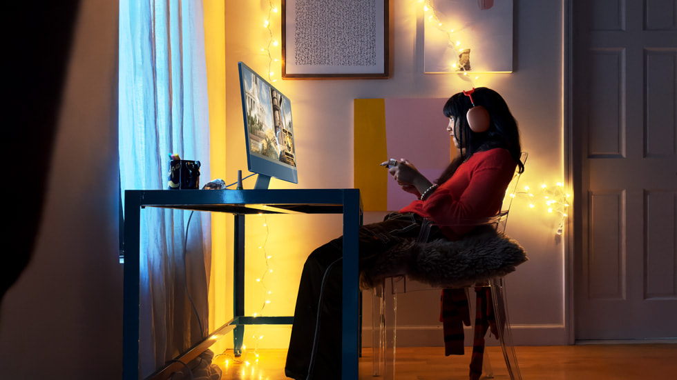 Seorang gamer terlihat sedang menggunakan iMac baru dengan M3 dan mengenakan AirPods Max berwarna merah.