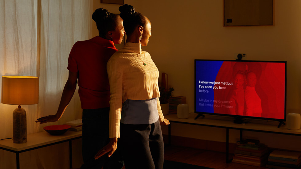 Dua pengguna Apple berpose saling memunggungi dan tercermin di layar TV mereka. 