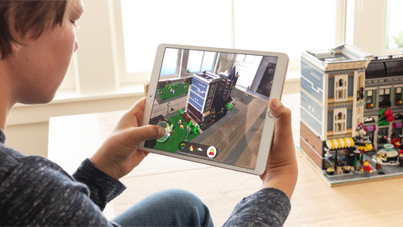 Bir odadaki LEGO binas?n?n ?nünde otururken, ekran?nda LEGO AR City olan iPad’i tutan bir erkek ?ocuk. 
