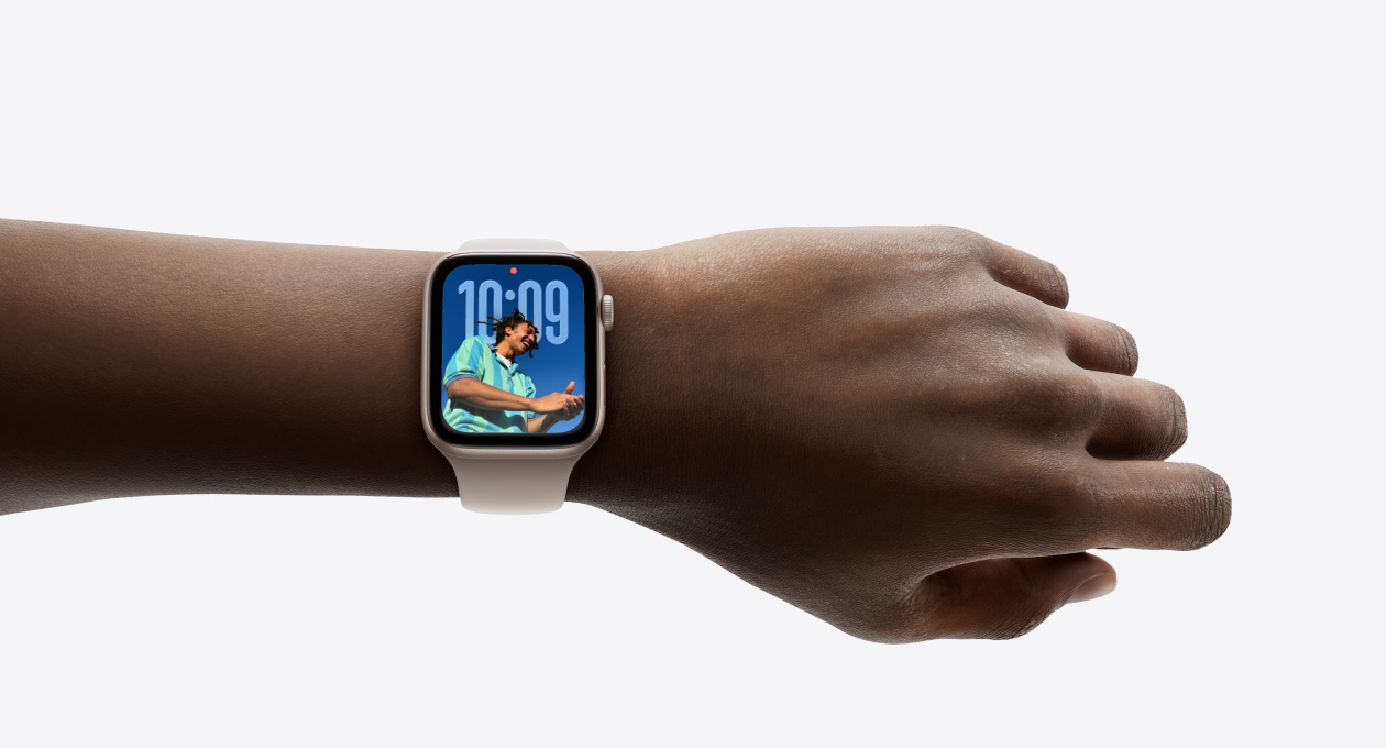 Primer plano de la mu?eca de una persona con un Apple Watch en color plata y correa de goma, en cuya pantalla se puede apreciar la app Hora en el nuevo dise?o con Liquid Glass