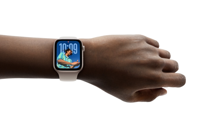 Primer plano de la mu?eca de una persona con un Apple Watch en color plata y correa de goma, cuya pantalla muestra la hora en el nuevo dise?o con Liquid Glass